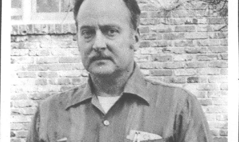 Black and white photo of balding man with mustache wearing striped collared button down shirt, standing against a brick building