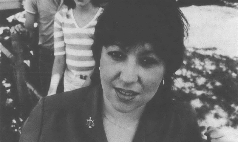 Black-and-white photograph of a woman with short hair, with two teenaged children standing behind her, looking directly at the camera and standing in front of a home.