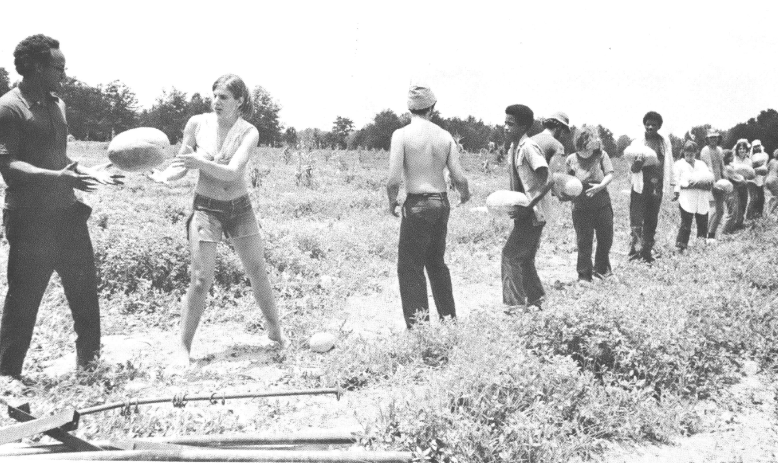 Black and white photograph of a line of young people, black and white, standing in a field passing watermelons in assembly-line fashion. 