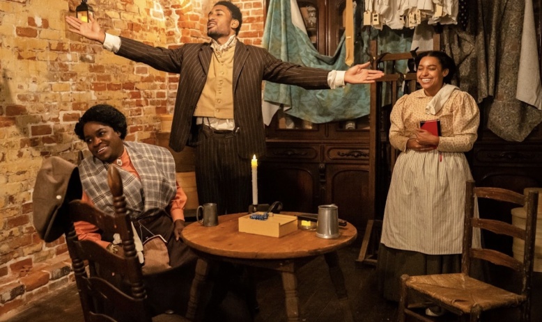 Three Black actors, two women and a man, wearing late 19th century clothing and gathered around a table