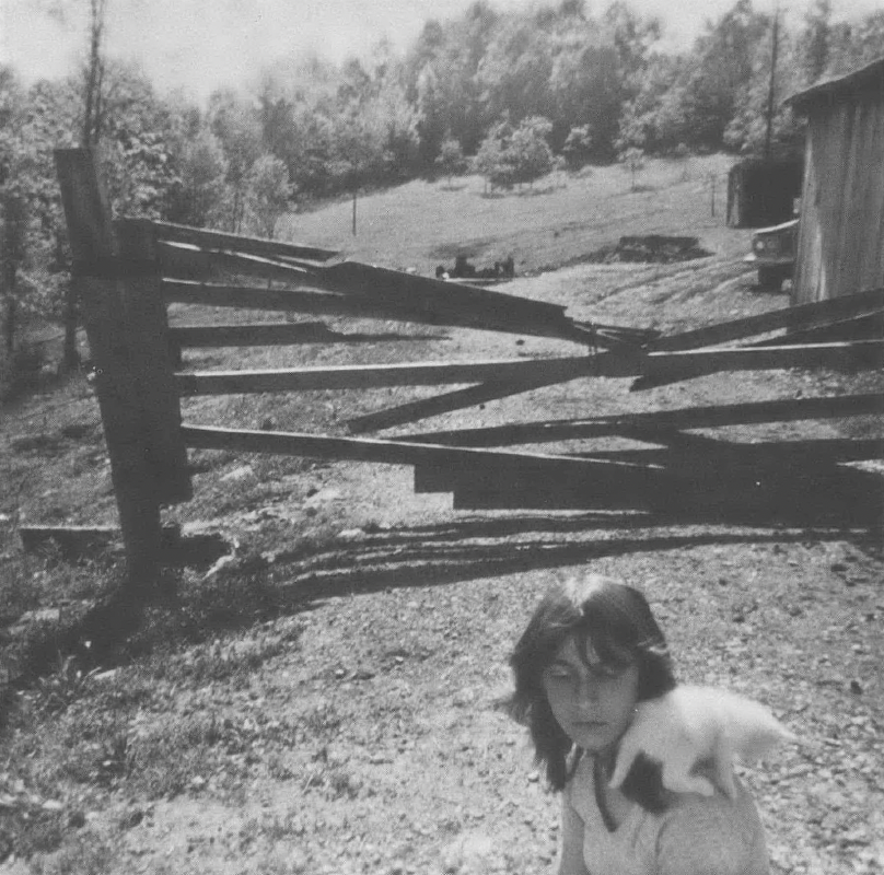 Black and white photo of girl with animal on her shoulder in front of falling-down fence