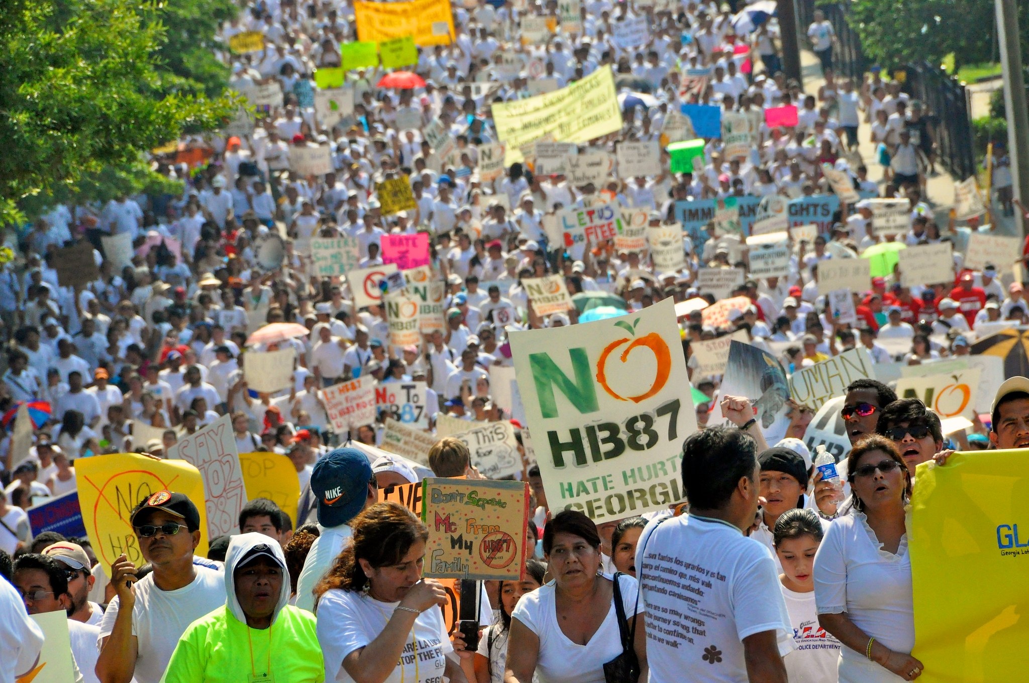 Image depicts a crowd of protesters carrying signs and marching in downtown Atlanta. In the foreground, a sign reading "No HB87, Hate Hurts Georgia" can be read.