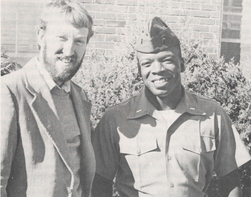 Black and white photo of two young men smiling at the camera, one white and one Black, dressed in military uniform