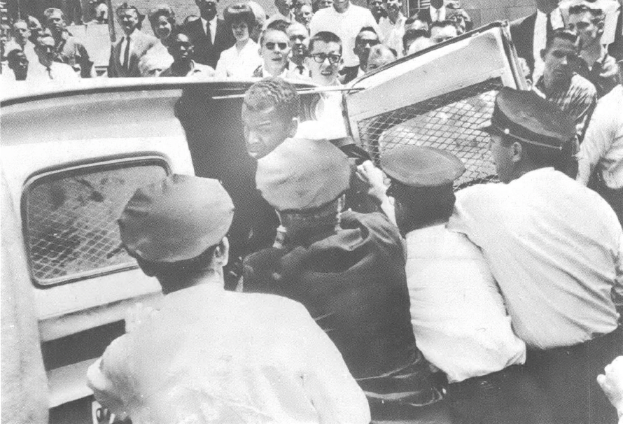 Black and white photo of police pushing people into car with mob around