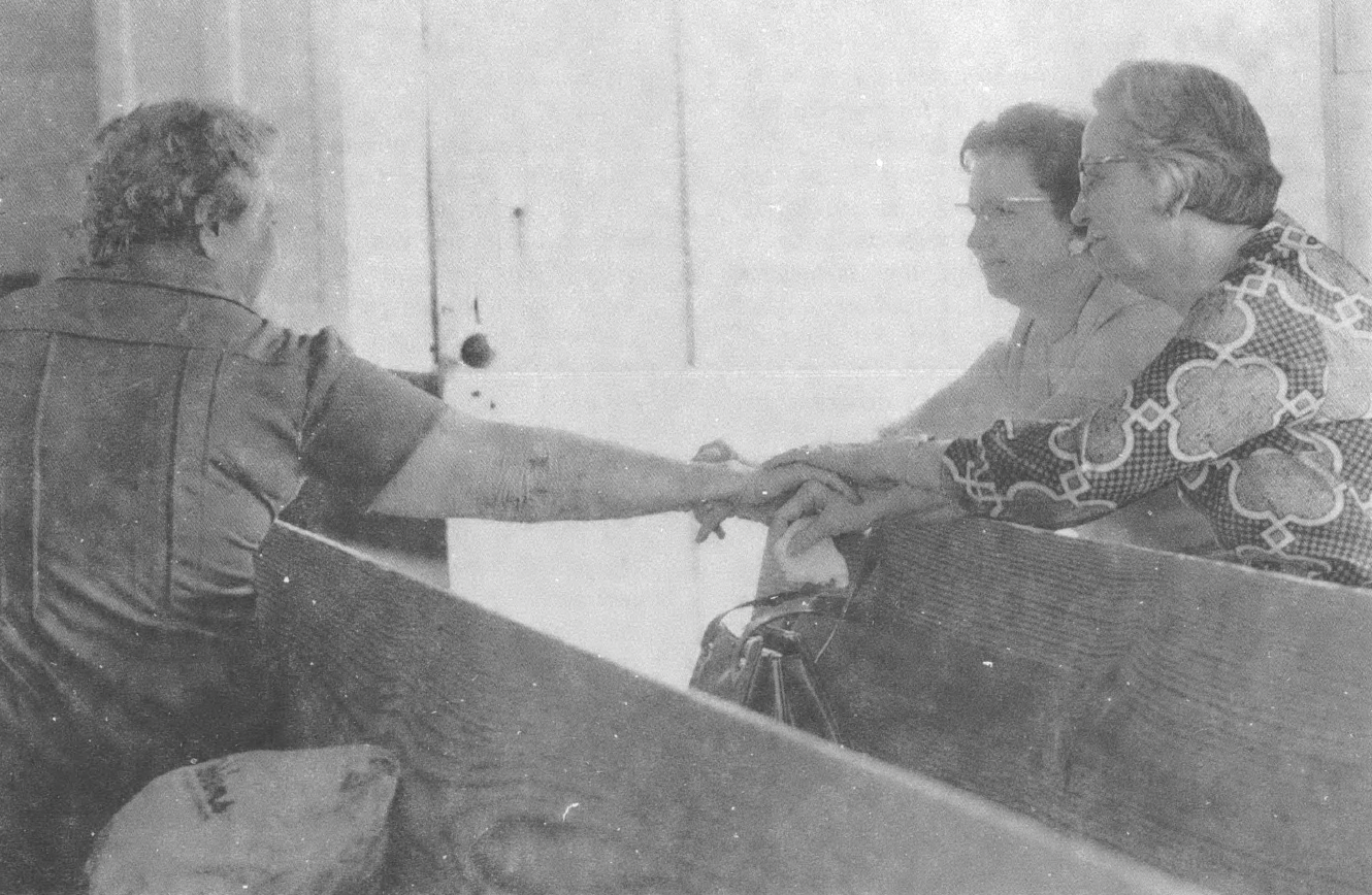 Black and white photo of three white women sitting in church pews, one woman shaking another woman's hand