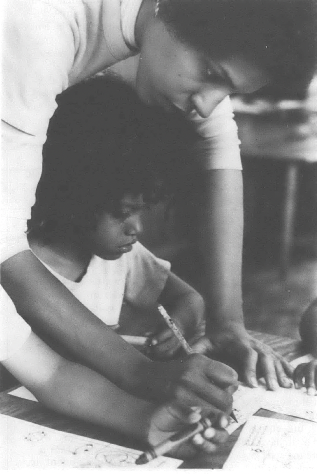 Black and white photo of Black woman helping Black child, seated at desk, write