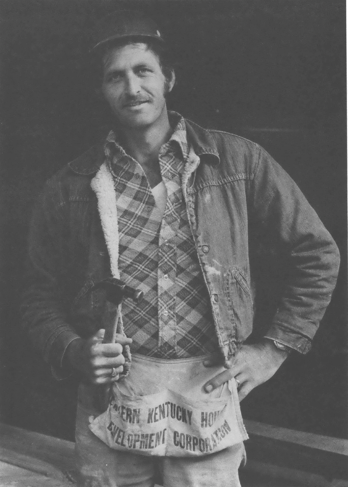 Black and white photo of young man wearing plaid shirt, jacket, and tool belt