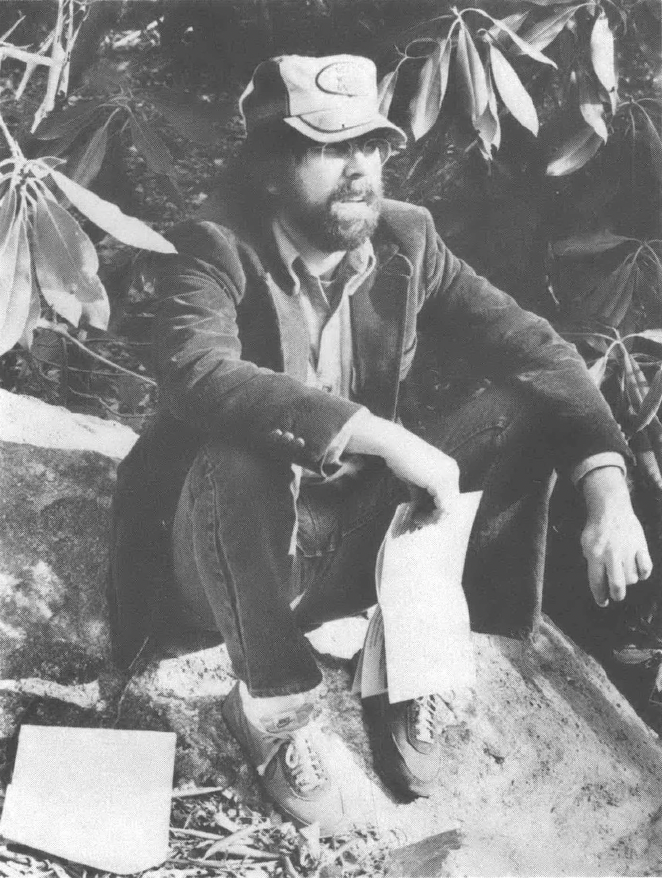 Black and white photo of young man wearing trucker hat holding papers sitting on the ground