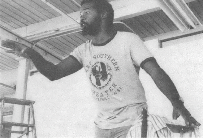 Black and white photo of Black man in t-shirt performing on stage, pointing at audience