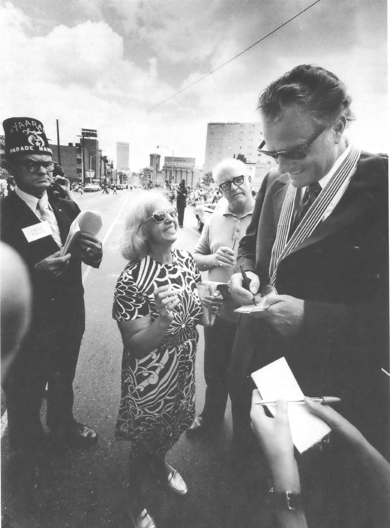 Black and white photo of middle-aged Billy Graham signing autographs surrounded by admirers 
