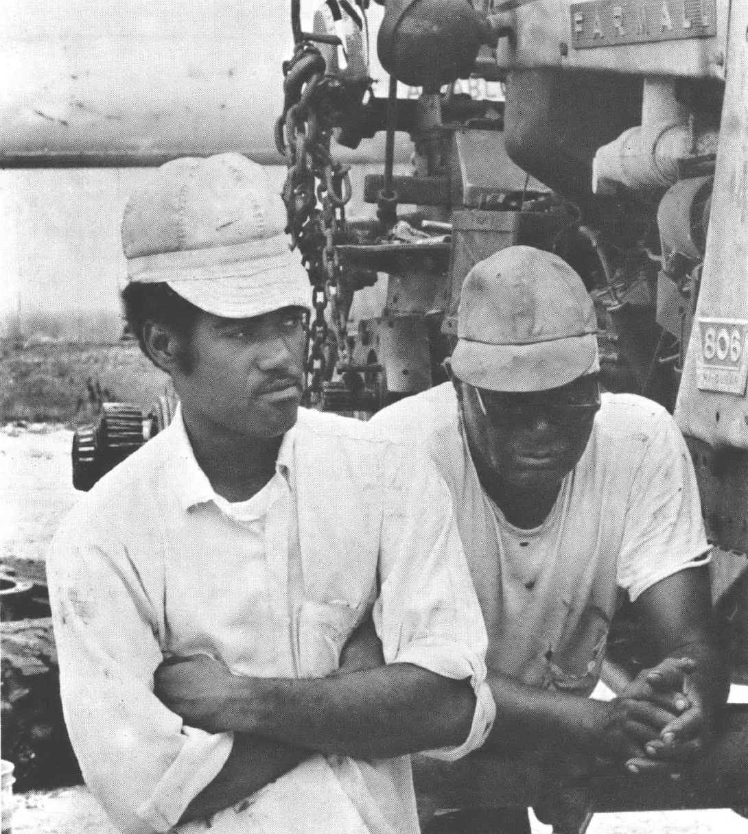 Black and white photo of two Black men, one younger and one older, both wearing trucker hats and standing next to a piece of agricultural equipment