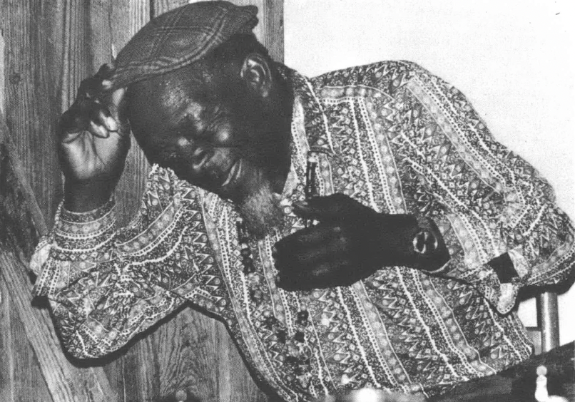 Black and white photo at an angle of Black man wearing patterned button-down shirt and cap, appears to be singing