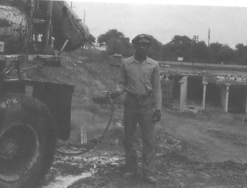 Black and white photo of Black man holding hose next to what looks like a cement mixer