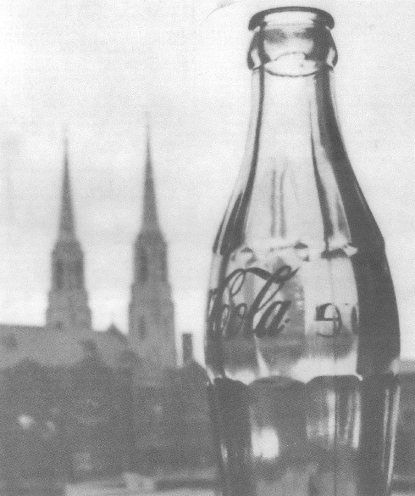 Black and white image of glass Coca-Cola bottle in front of church with two steeples