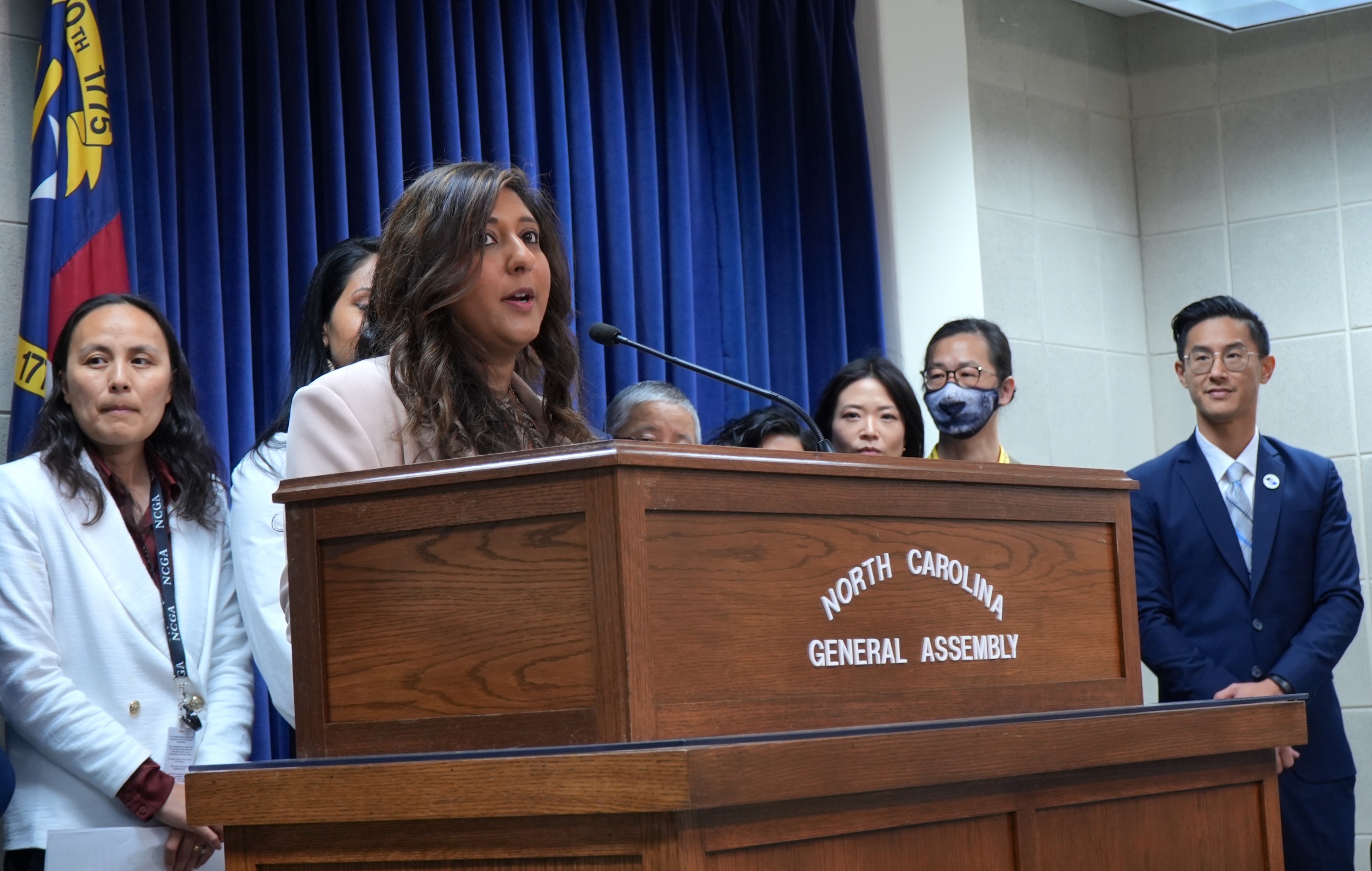 An Indian American woman speaks at a wooden podium that has the words "North Carolina General Assembly" attached to the front. Several other people who are Asian American stand behind the woman, watching her speak.