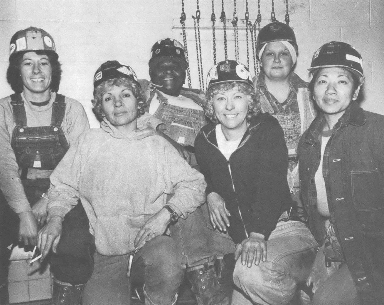 Black and white photo of six women of different races and ethnicities in coalmining gear