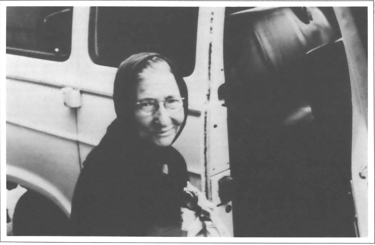 Photo of older woman with scarf around her head next to a car