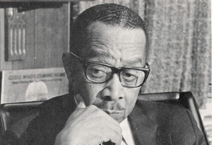 Black and white portrait of Black man with glasses, in suit, and hand on chin posed in front of bookcase