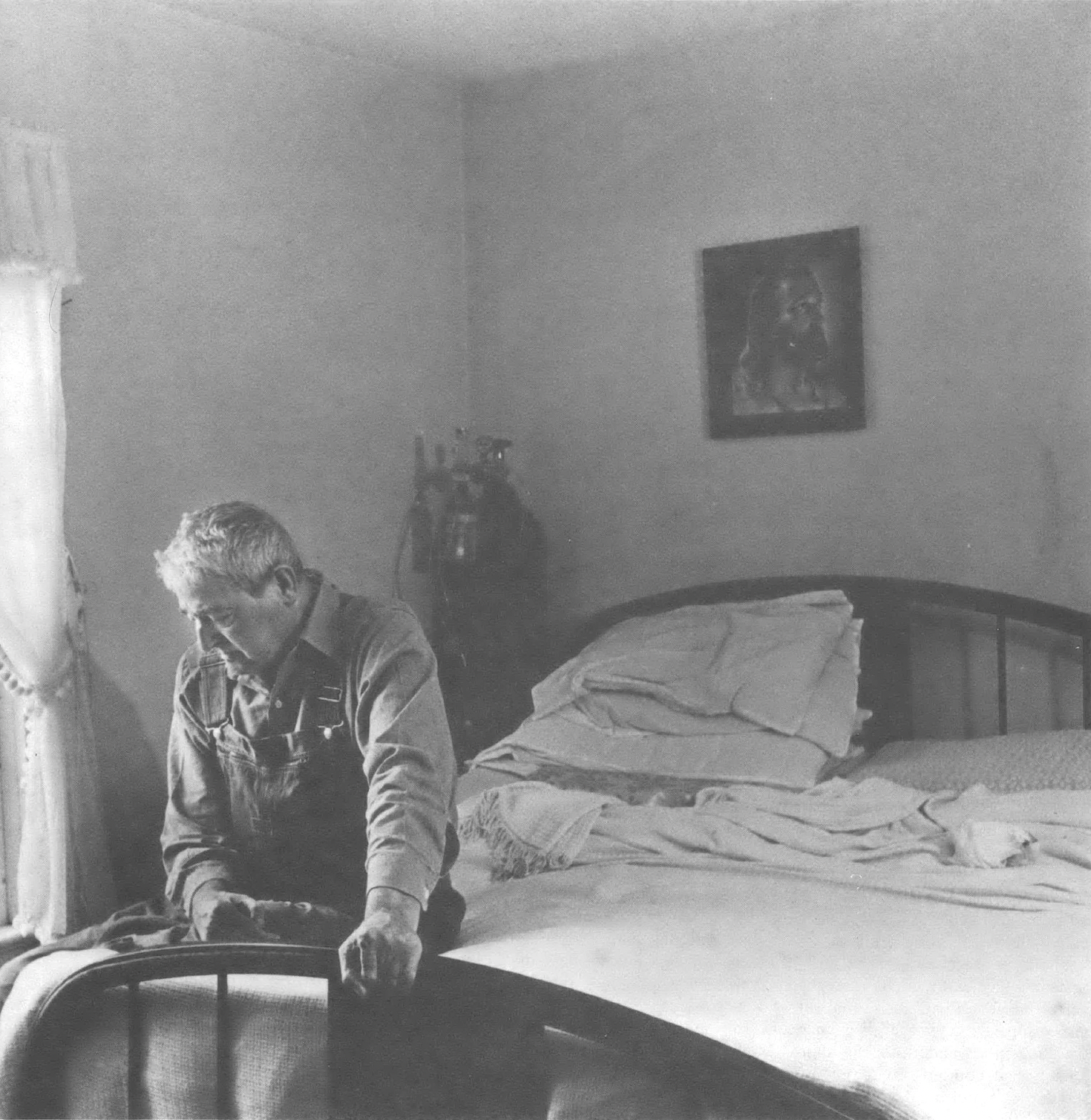 Black and white photo of elderly white man sitting at the end of a bed looking down at the floor