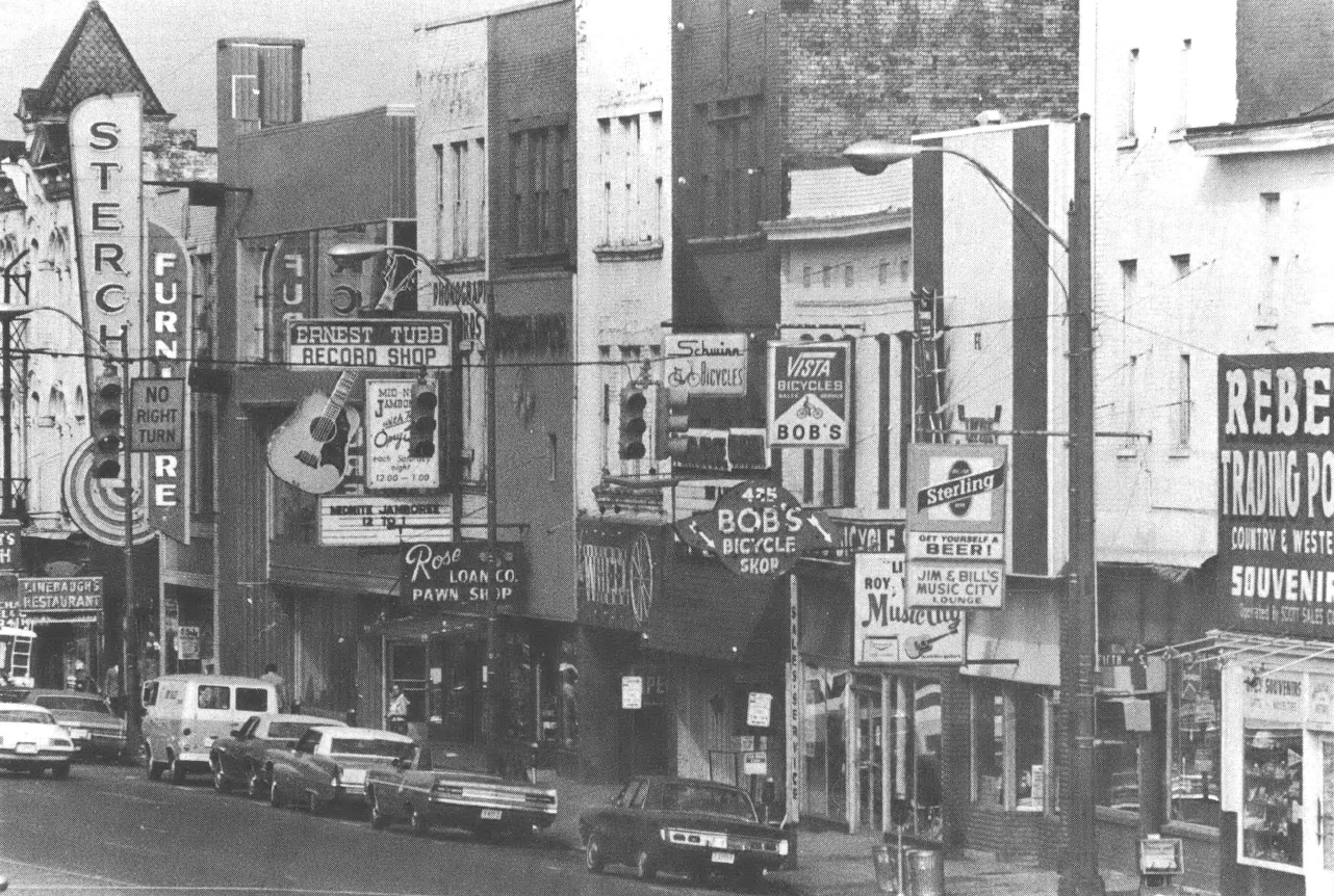 Black and white photo of city street with several record and music shops, and cars parked on the street in front