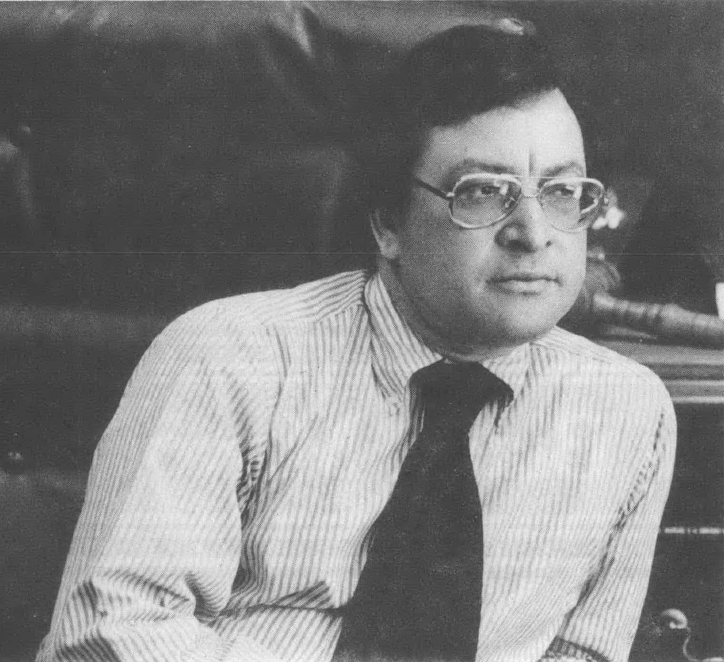 Black and white photo of Dutch Morial. wearing eyeglass, a button-down shirt, and tie, sitting and leaning to his left