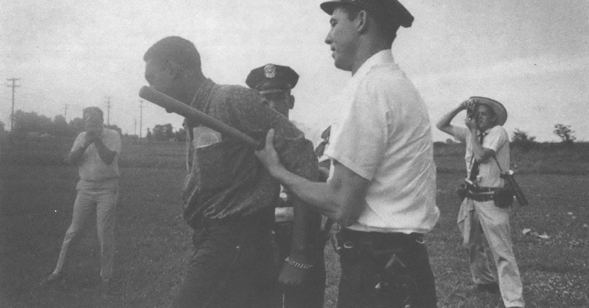 Black and white landscape photo of cops arresting a Black man in a field, with photographers behind
