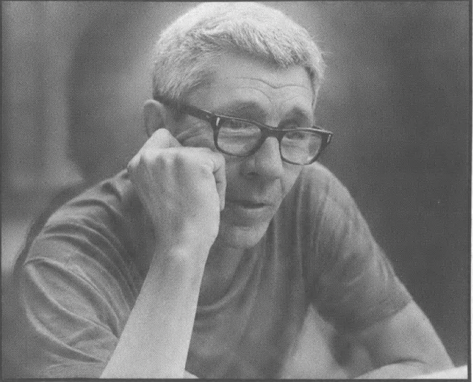 Black and white image of white man with grey/white hair and glasses, head resting on one hand