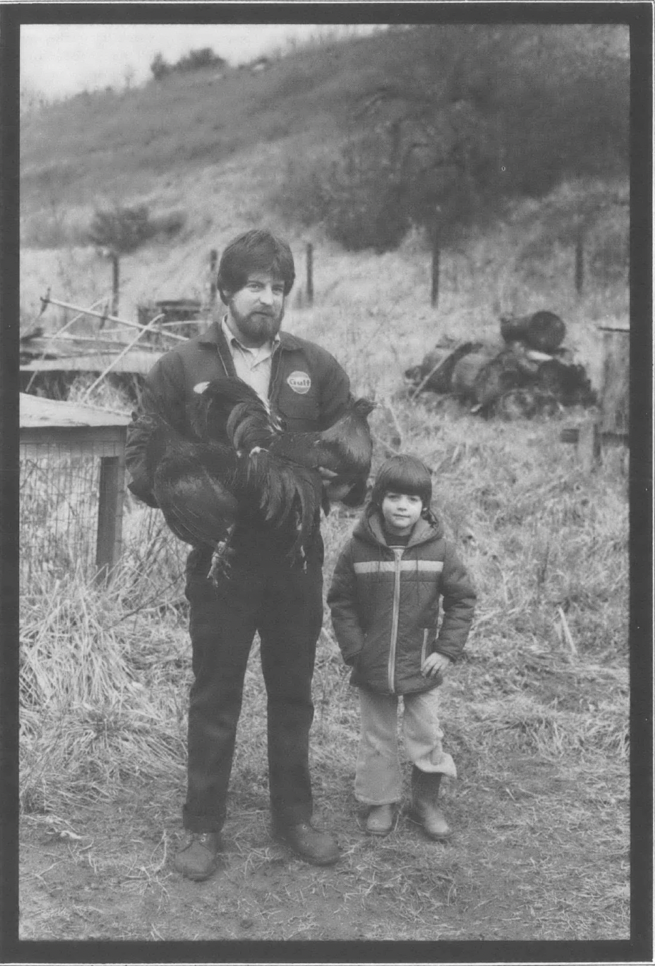 Black and white photo of white man and child standing outside against farm backdrop, wearing coats