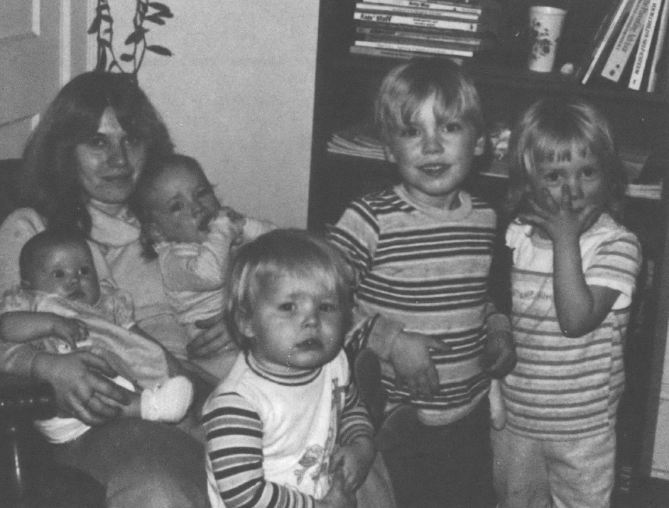 Black and white photo of seated white woman holding two babies with three other young children standing in front of her