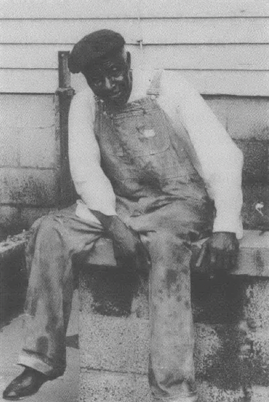 Black and white photo of older Black man in overalls and hat sitting on a bench outside of a building, slightly hunched over