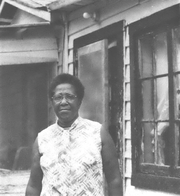 Black and white photo of middle-aged Black woman standing in front of a home