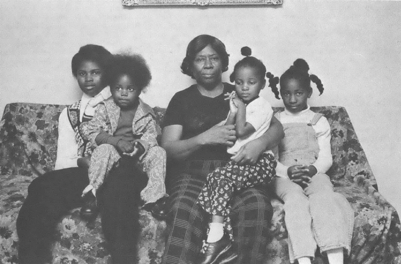 Black and white family photo of Black woman with four young children sitting on a couch inside a home. 