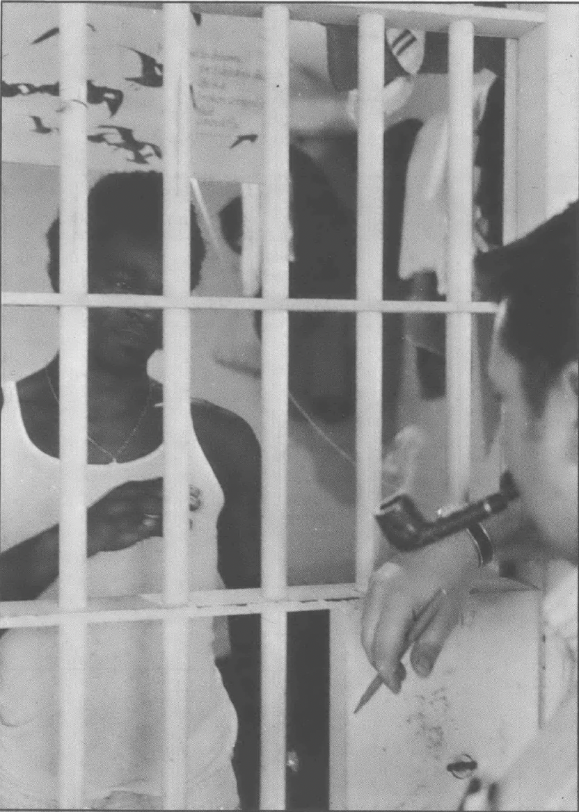 Black and white photo of a white man smoking a pipe facing a Black man who is in a cell