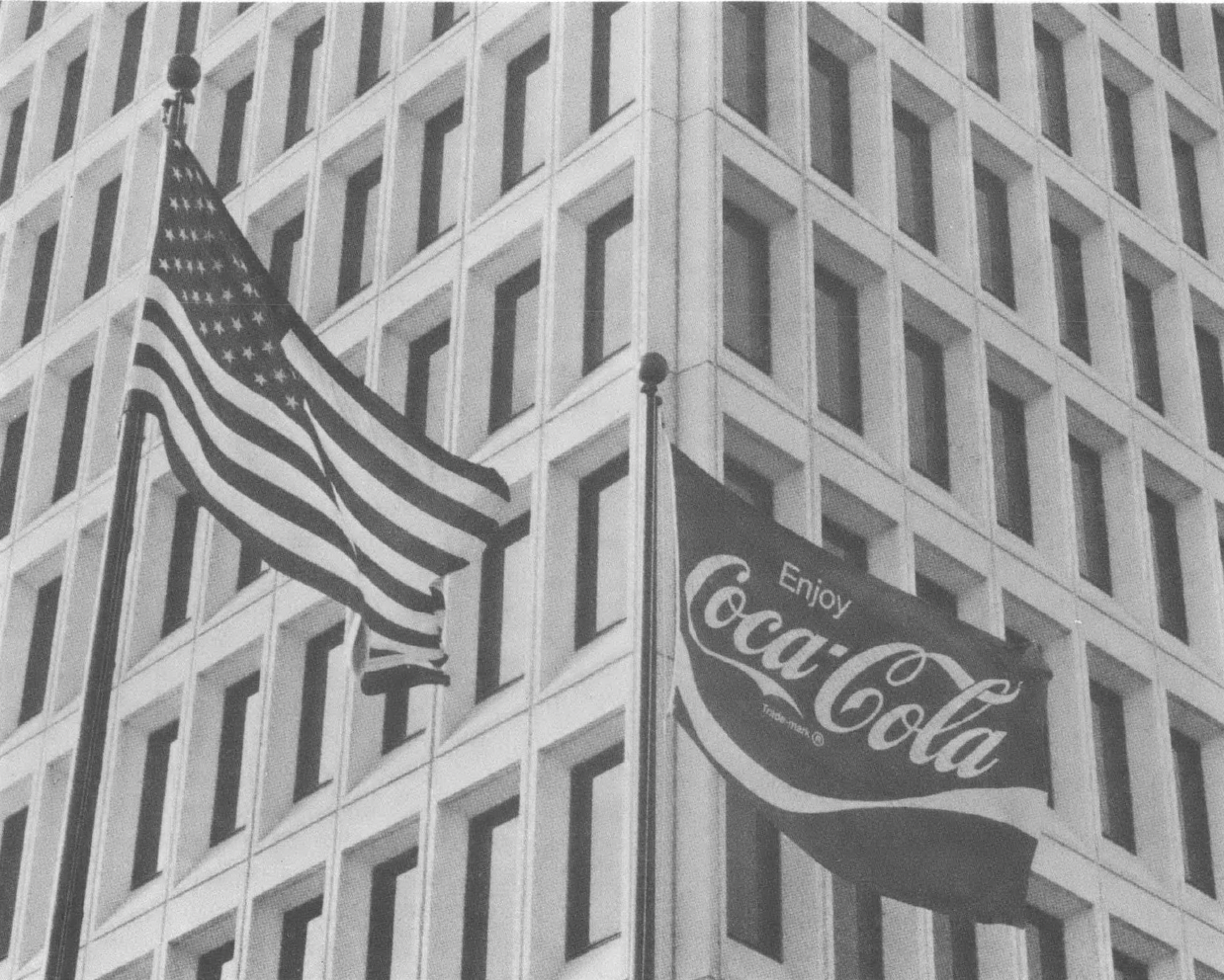 Black and white photo of American flag and Coca-Cola flag flying in front of high-rise building