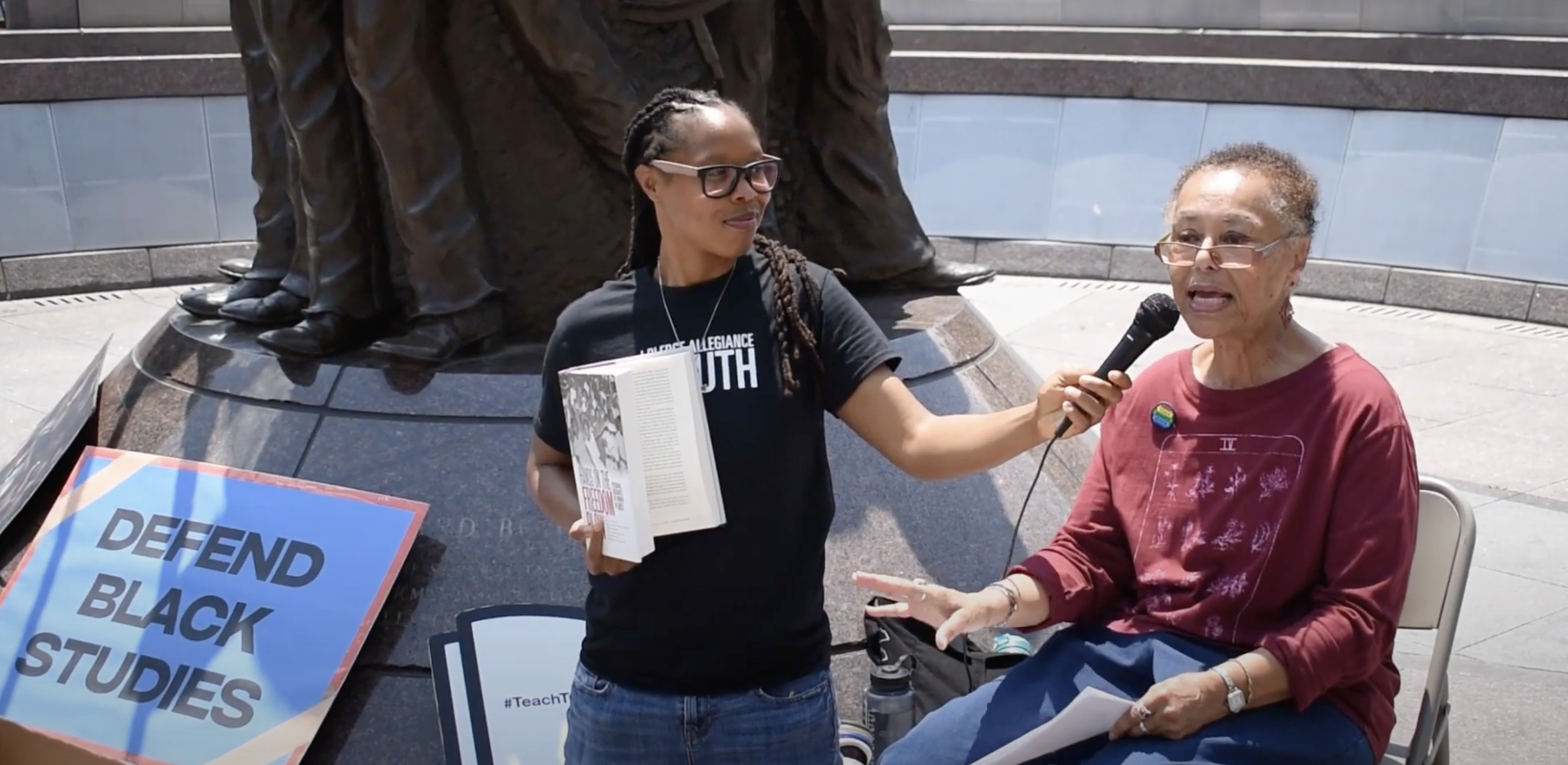 A Black woman wearing a black t-shirt holds out a microphone for another older Black woman who is seated and wearing a red t-shirt to speak into. They are at the African American Civil War Memorial in Washington, D.C. A sign that reads "Defend Black Studies" lays in the corner.