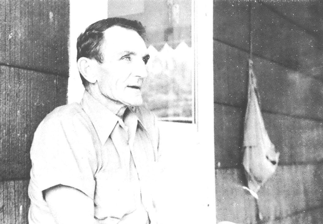 Black and white photo of older white man seated outside in front of the window of a house, talking and looking at someone away from the camera