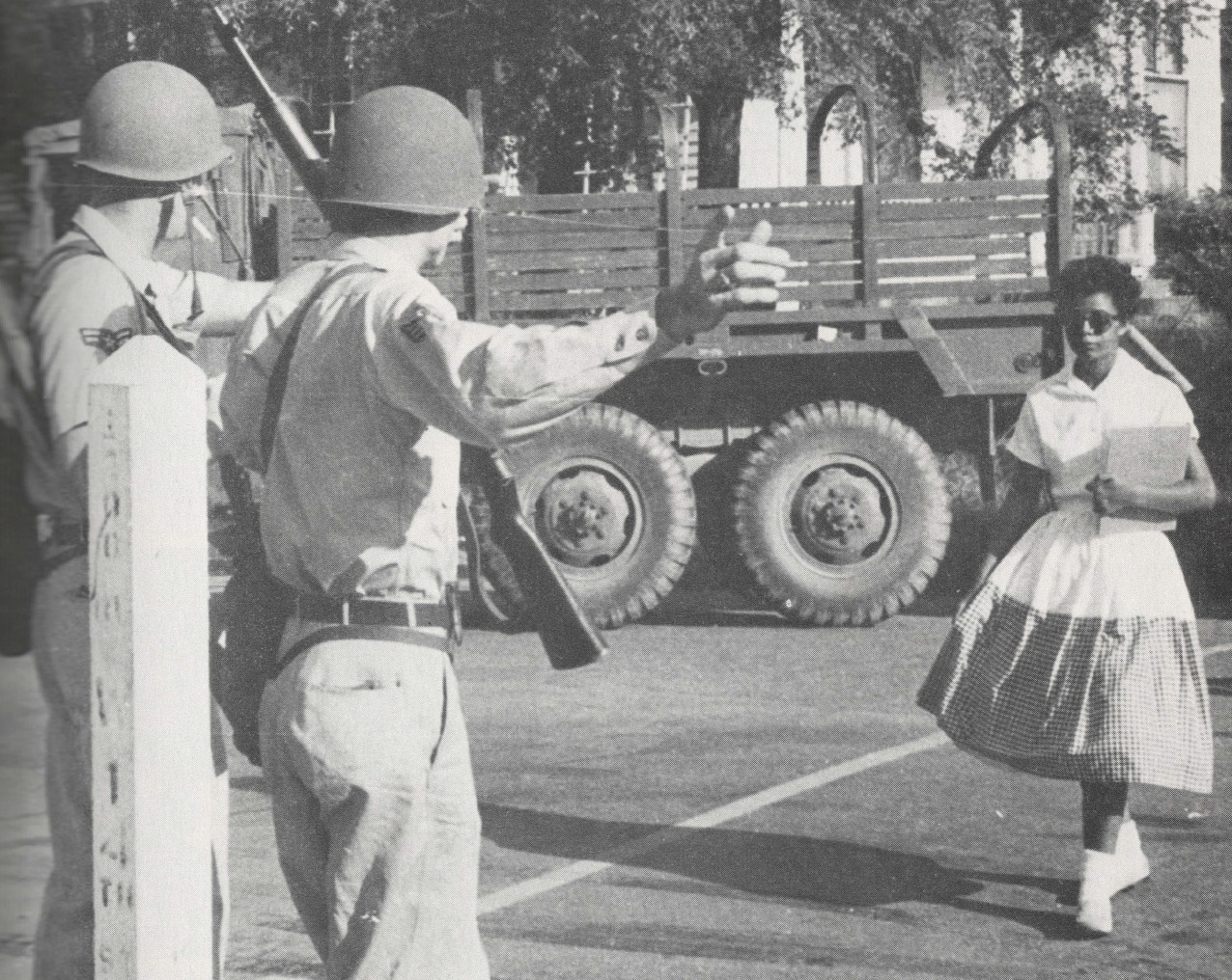 Black and white photo of young Black student in dress and sunglasses walking towards two National Guard members