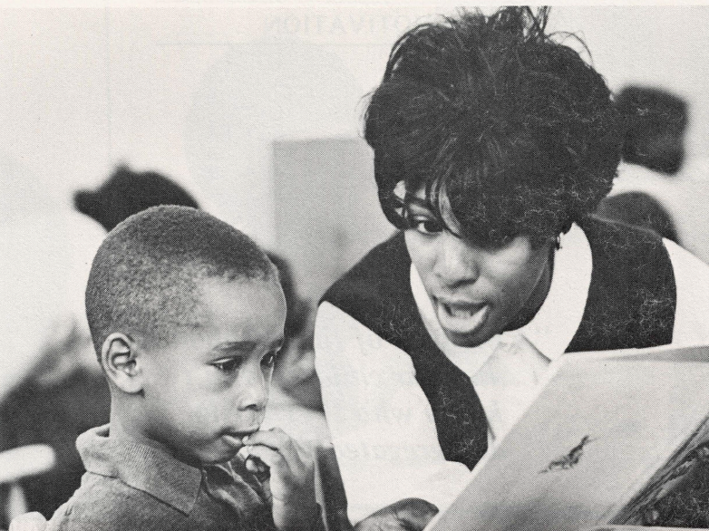 Black and white photo of Black teacher explaining a paper to young Black student in classroom