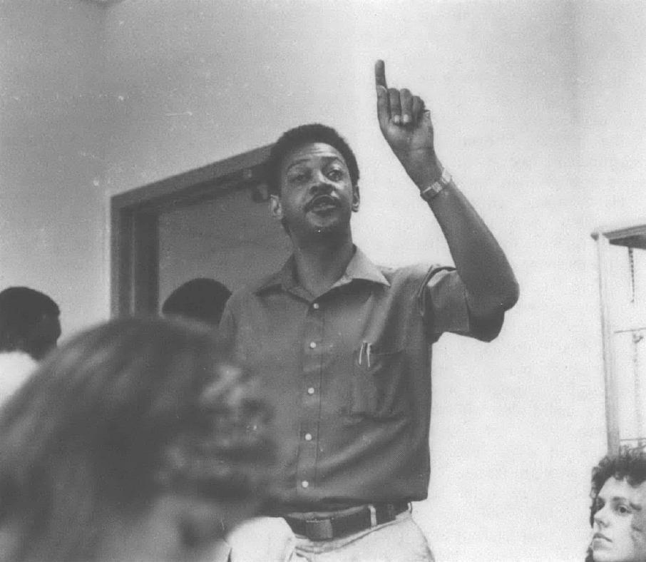 Black and white photo of Black man speaking to group of people in a room