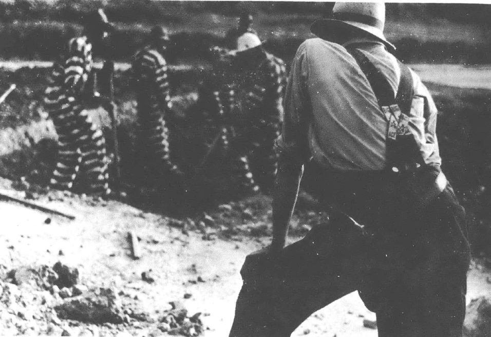 Black and white photo of a man with his back turned to the camera watching prisoners in black and white jumpsuits digging a hole.