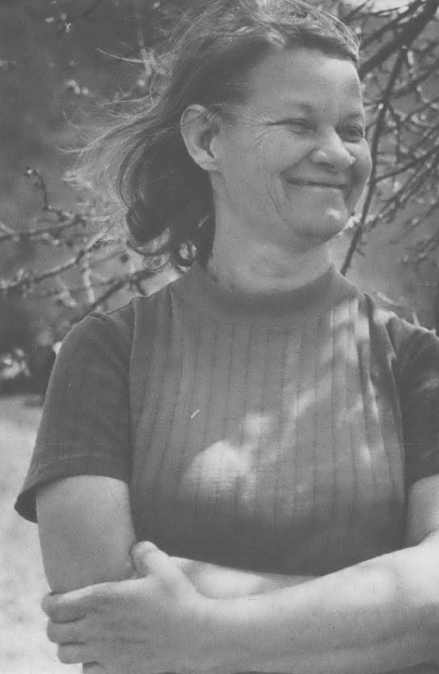 Black and white photo of white woman with her arms crossed smiling away from the camera, standing in a patch of sunlight