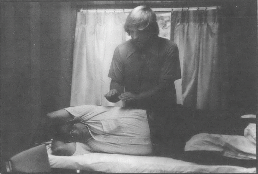 Black and white photo of man, standing, holding his hands over another man lying on his side on a table