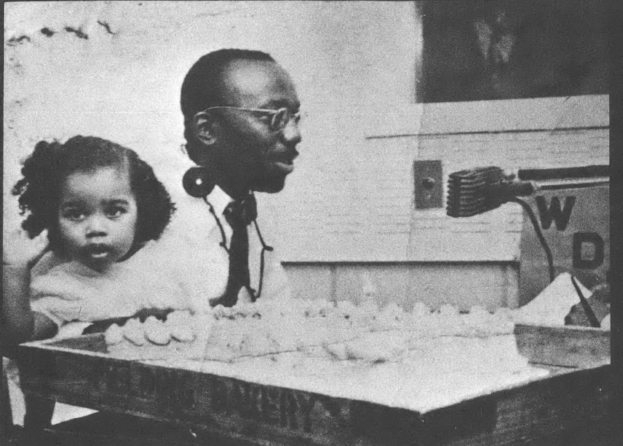 Black and white photo of Black man with glasses seated at table speaking into microphone, with toddler-age child seated next to him looking away from the camera