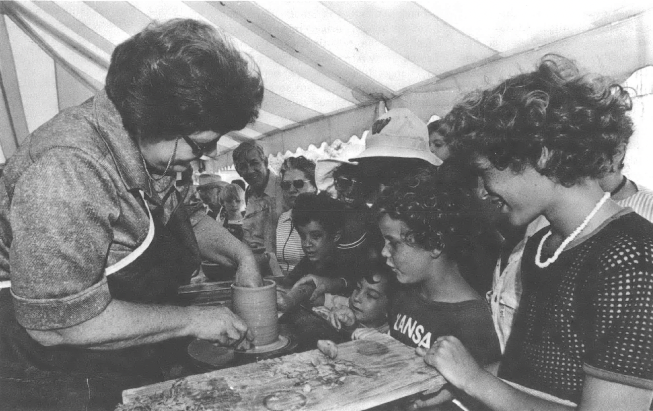 Black and white photo of older white woman showing a piece of pottery to children at a fair booth