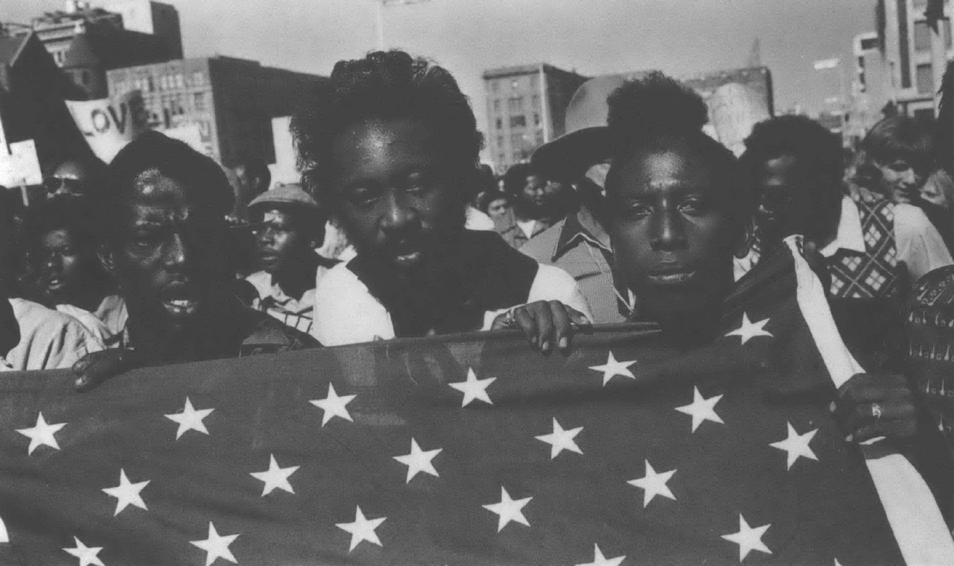 Black and white photo of Black activists marching, yelling while holding American flag