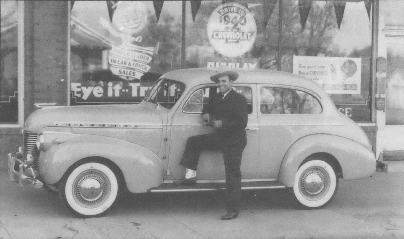 Black and white photo of man standing next to nice car in front of shop window