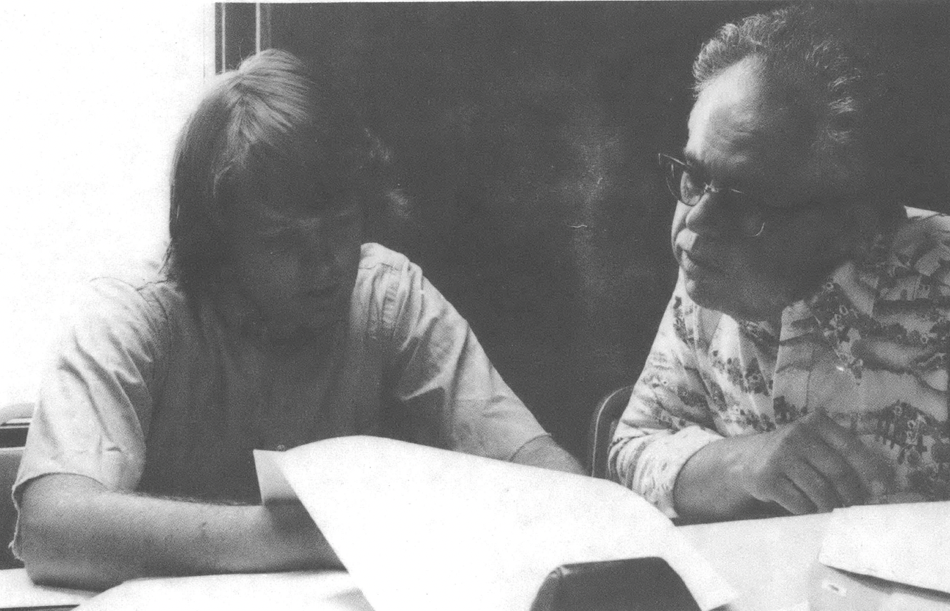 Black and white photo of two white men, one younger and one older, looking through papers at a table