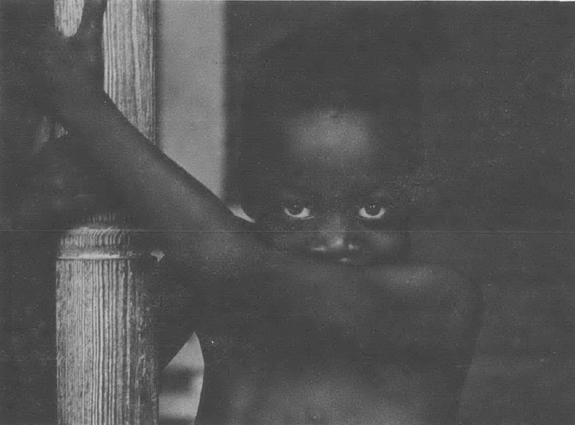 Shirtless Black child holding on to a wooden post and peering over their arm