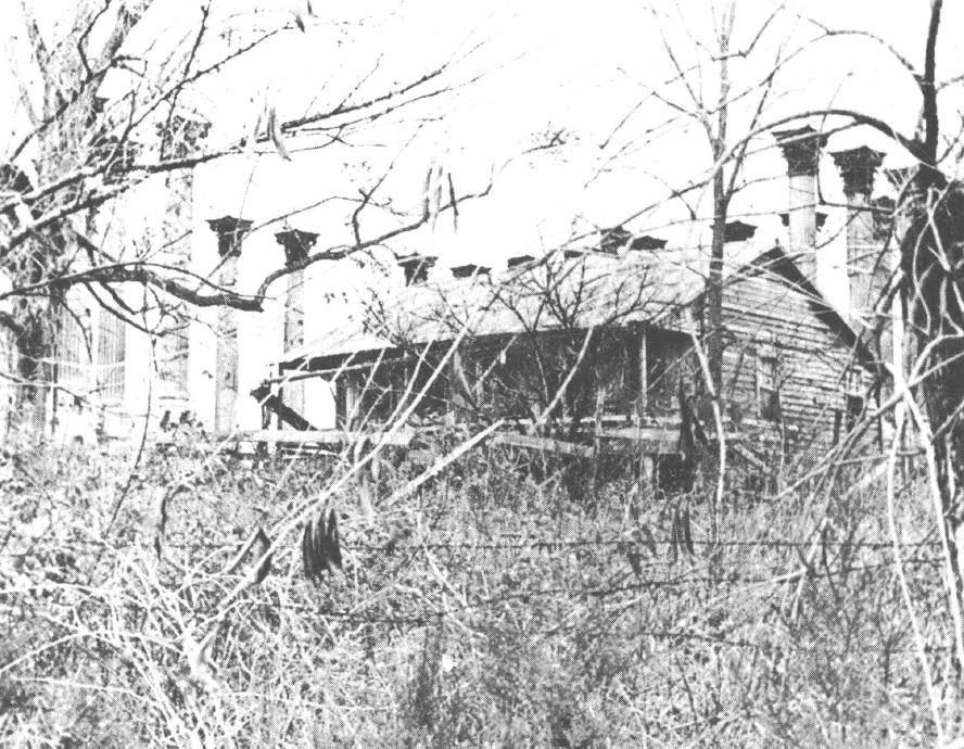 Black and white photo of building behind a barbed wire fence and tall grass