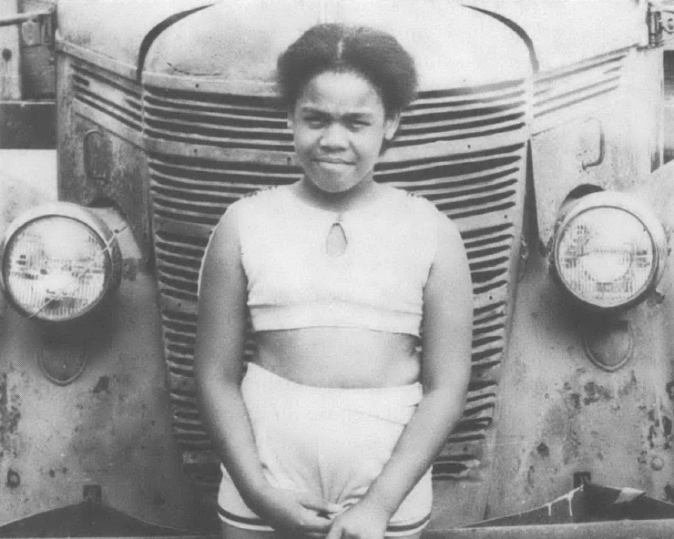 Black and white photo of Black child standing in front of old car
